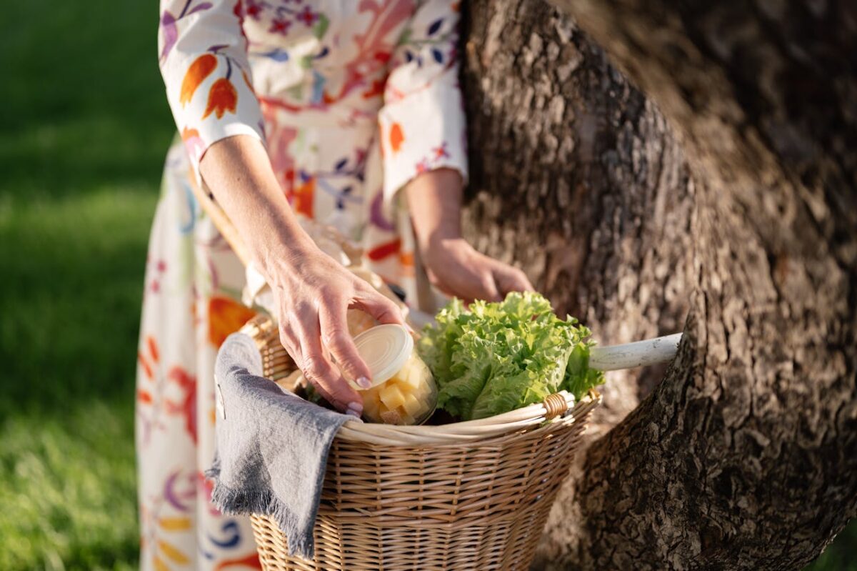 A woman in a floral dress places a jar in a wicker basket outdoors. Fresh produce visible.